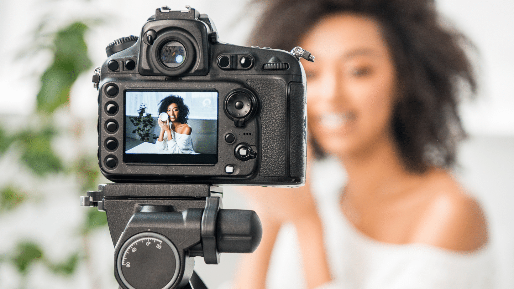 A person sitting in front of a video camera on a tripod filming a commercial for a beauty product.