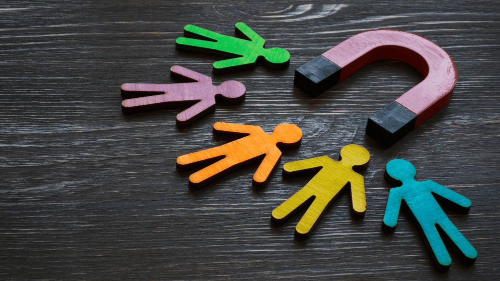 An Ascend Adwerks' image of multicolored individuals standing around a magnet on a wooden table representing Paid Social Media Ads Services.