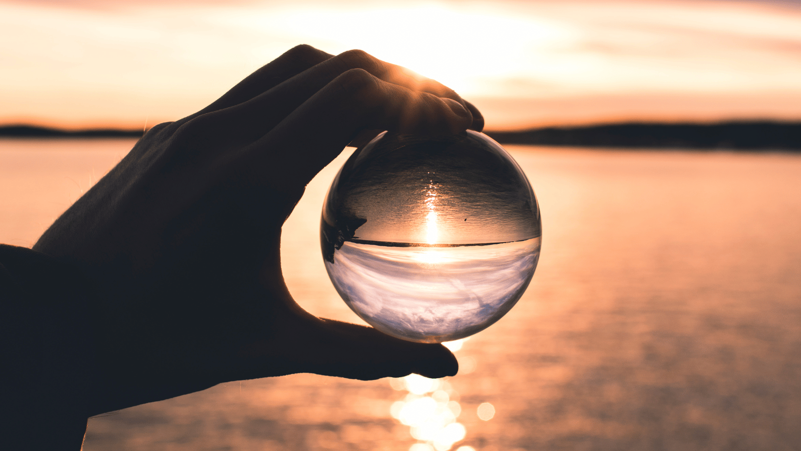 A person holding up a glass ball with the sun setting in the background.